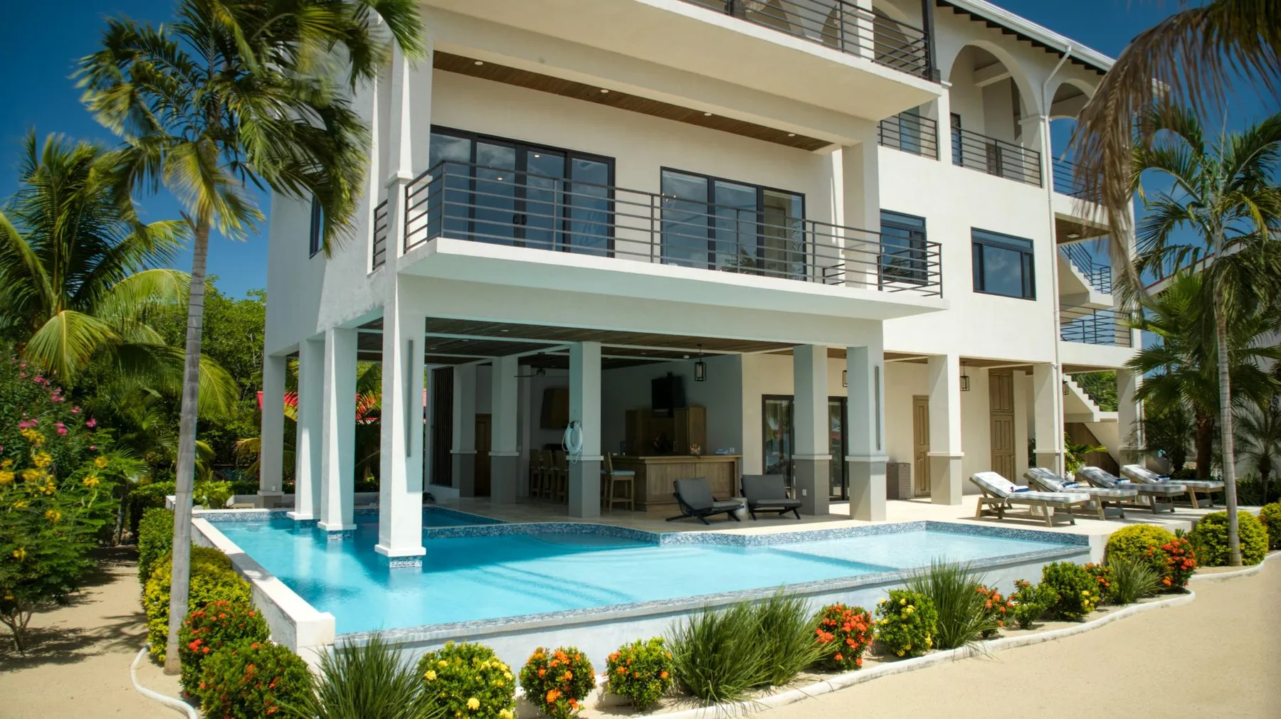A white multi-story building overlooks a swimming pool surrounded by tropical plants.