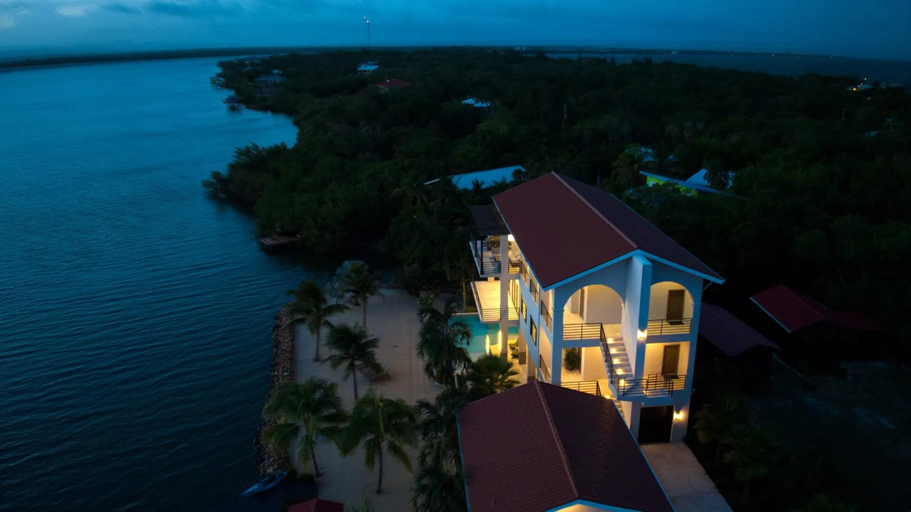 A lit waterfront building stands beside calm blue water and palm trees at dusk.
