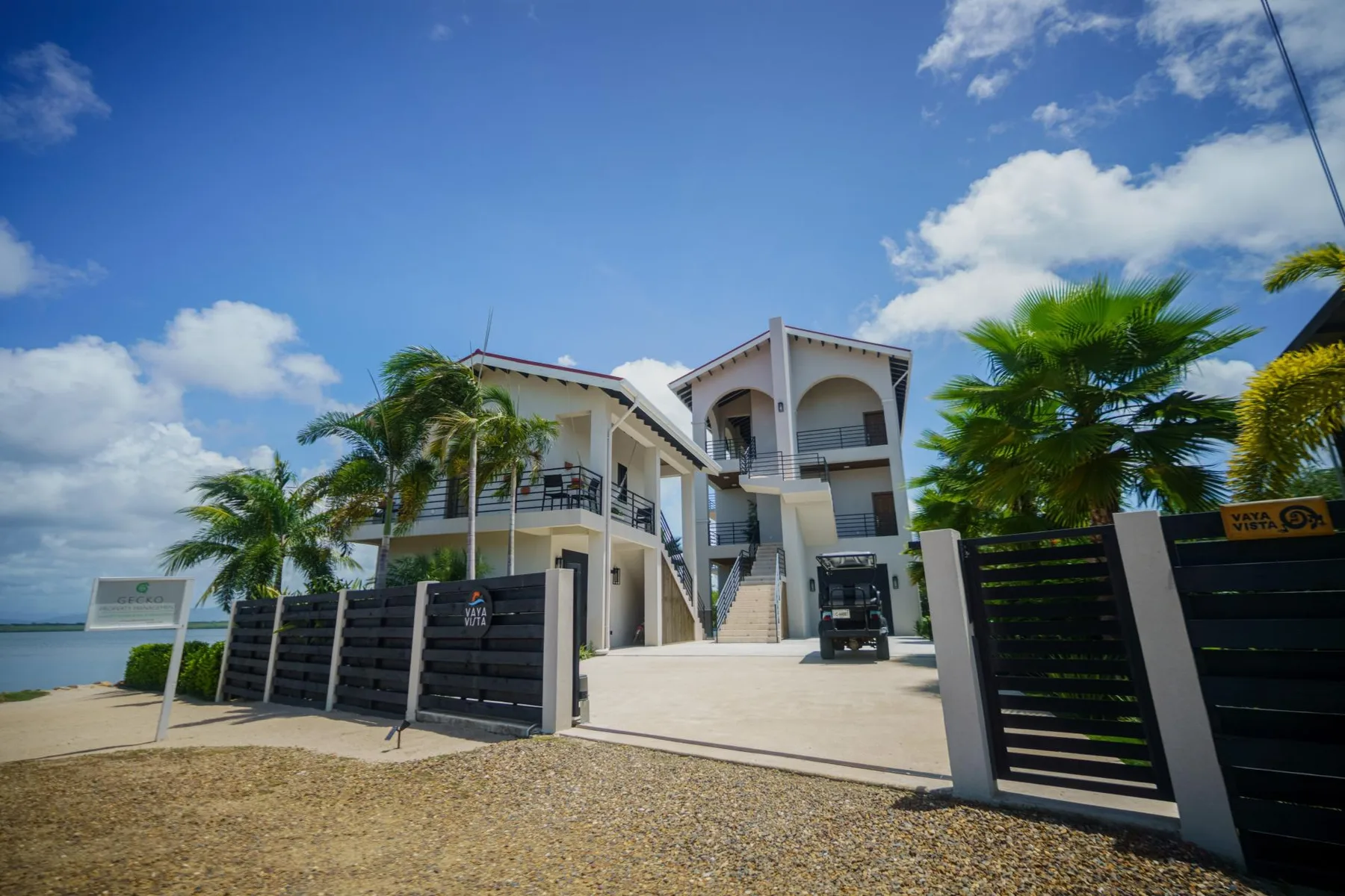 A white multi-level building stands behind a dark entrance gate, with palm trees nearby.