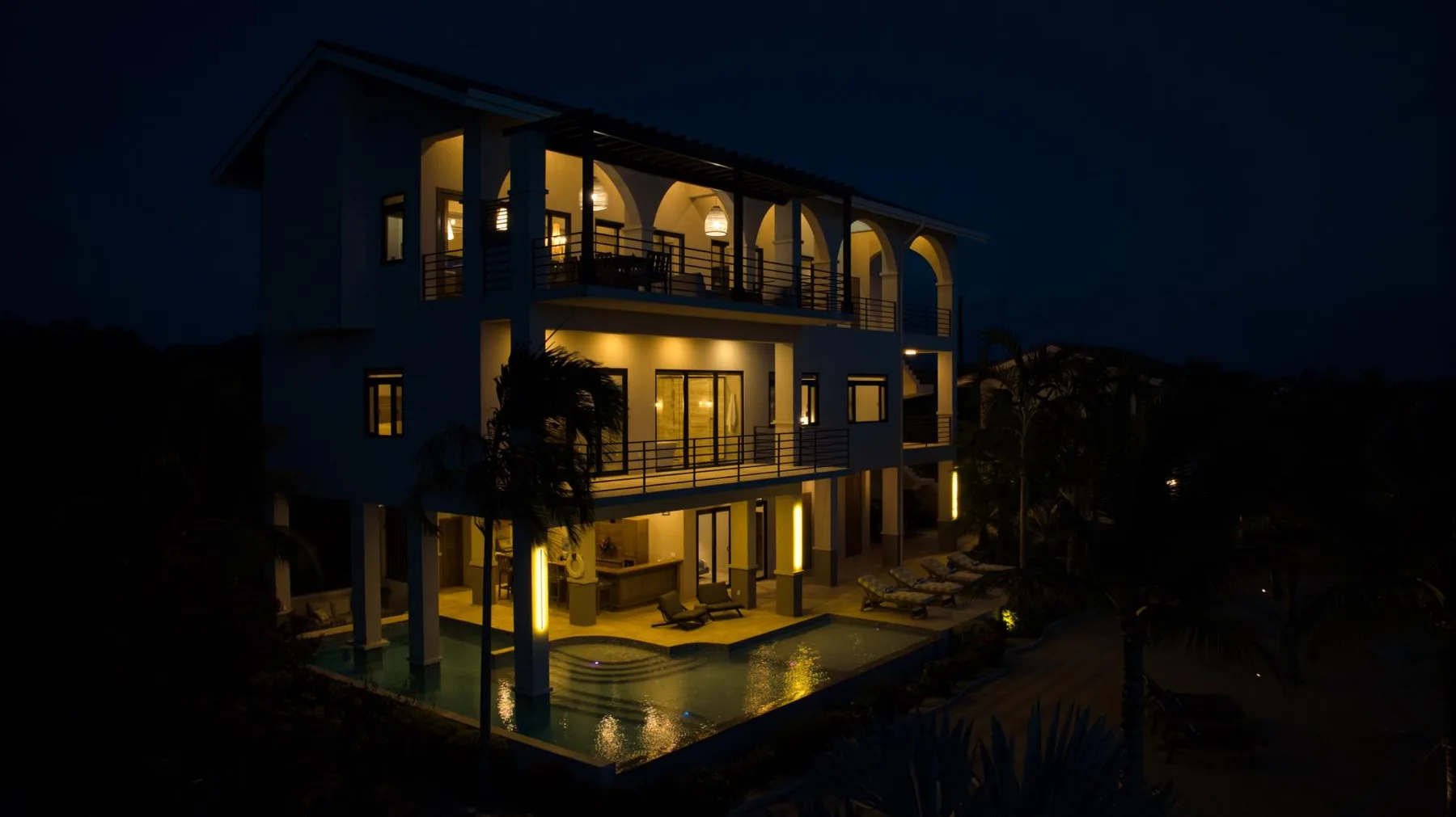A multi-level building glows with warm lights beside an illuminated swimming pool at night.