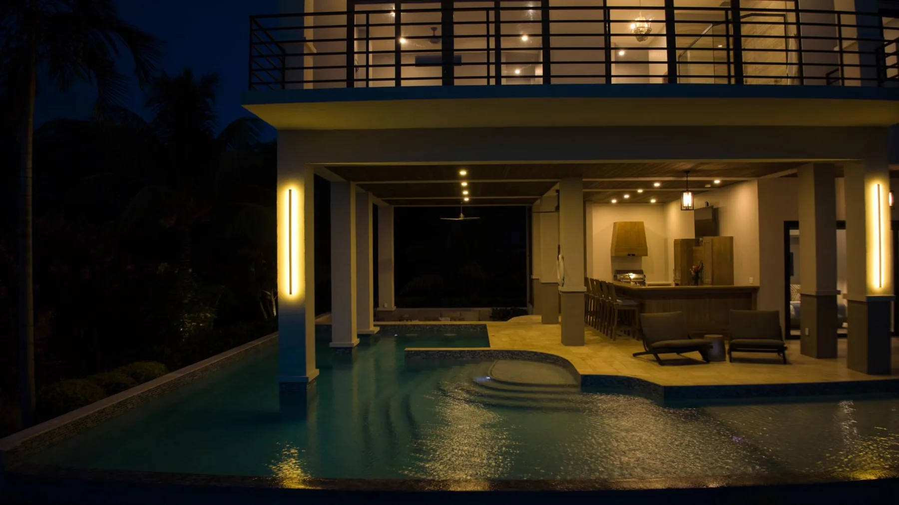 A lit modern house overlooks a swimming pool at night.