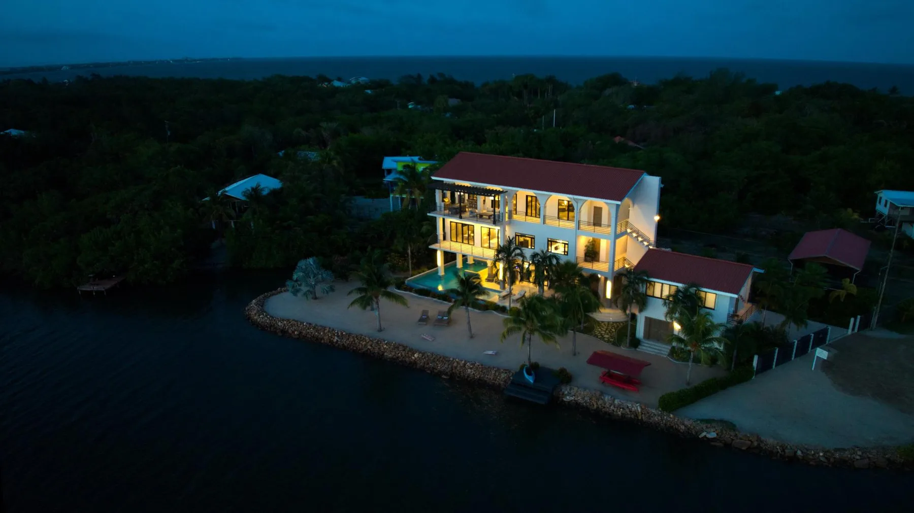 An illuminated waterfront building stands beside a tree-lined shore at dusk.