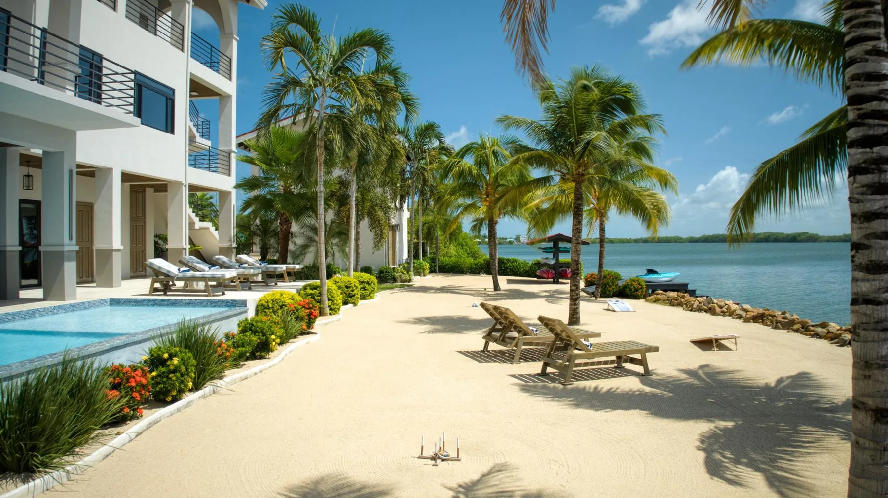 A sandy beachfront patio with lounge seating beside a pool and palm trees.