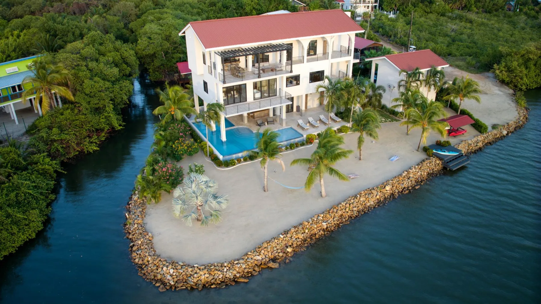A waterfront building with a red roof stands beside a pool and palm trees.