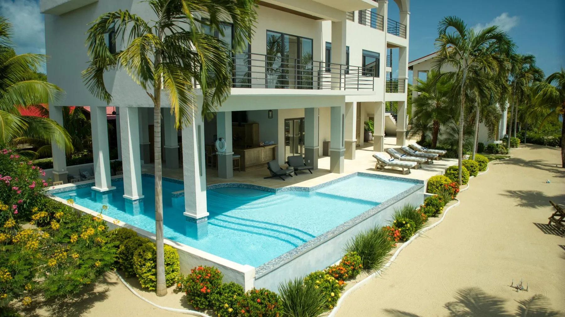 A modern building beside a swimming pool, with palm trees and lounge seating.