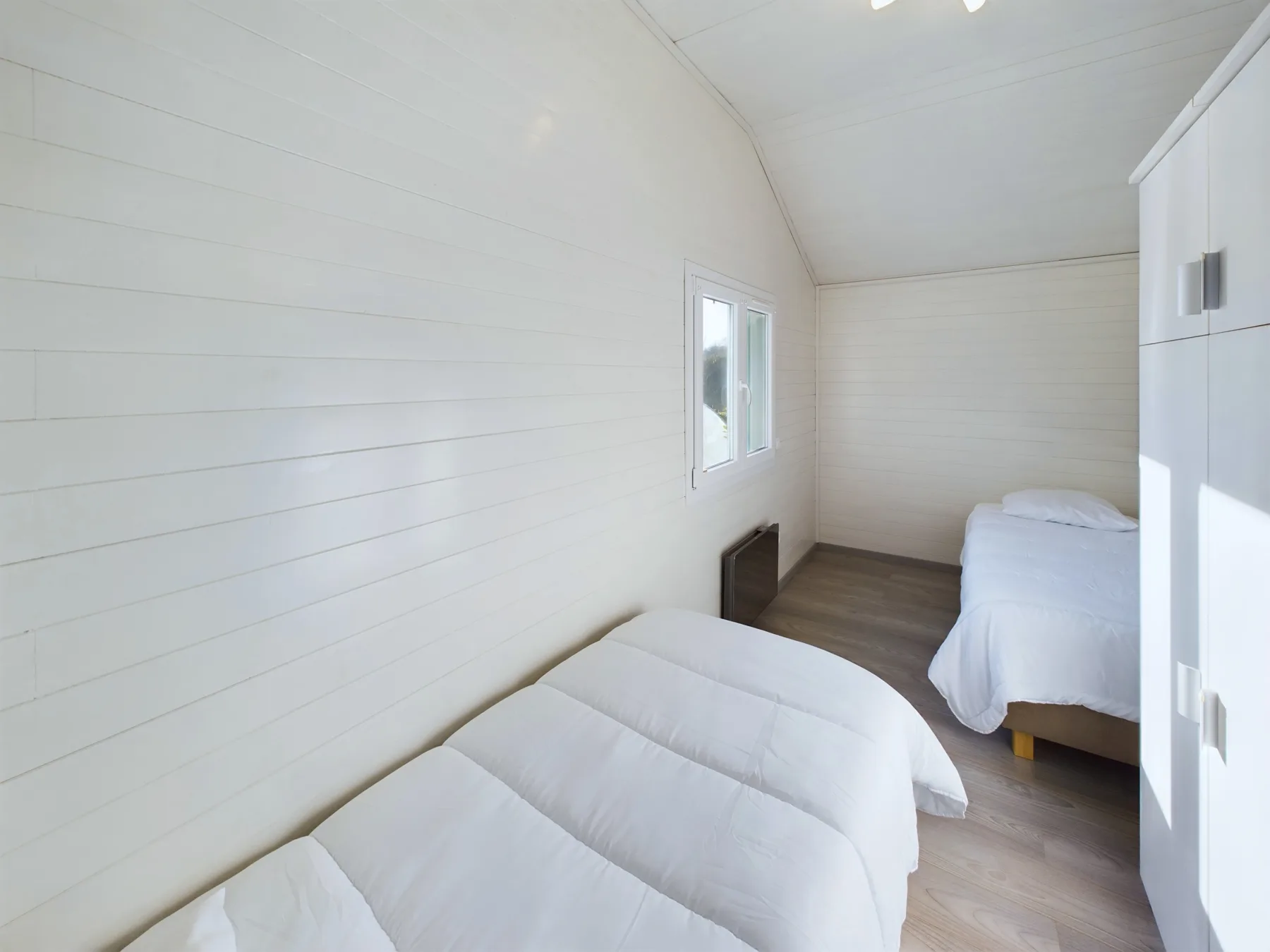A bright bedroom with two beds and a window along the paneled wall.