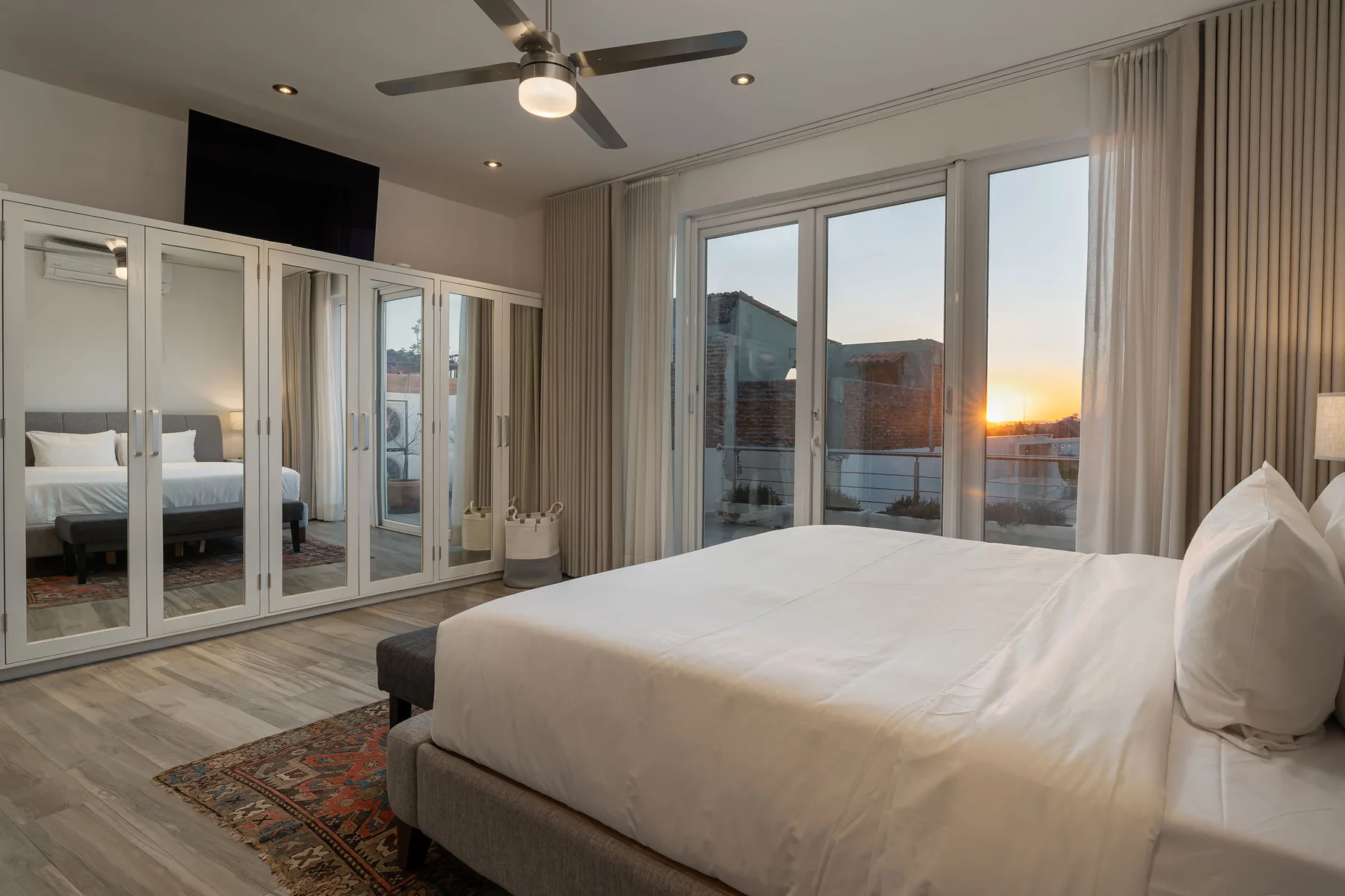 A bedroom with a large bed beside floor-to-ceiling windows overlooking the sunset.