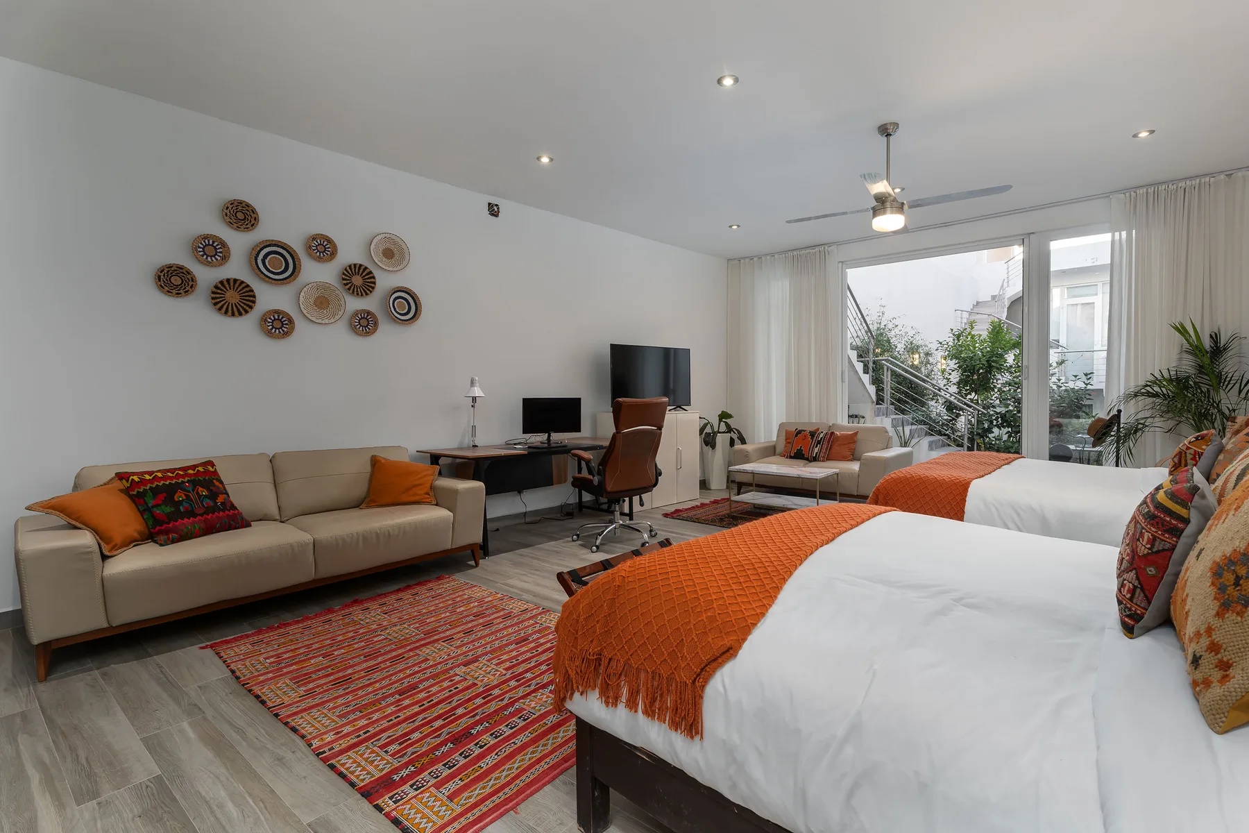A bright bedroom with beds, a seating area, and a desk beside large windows.