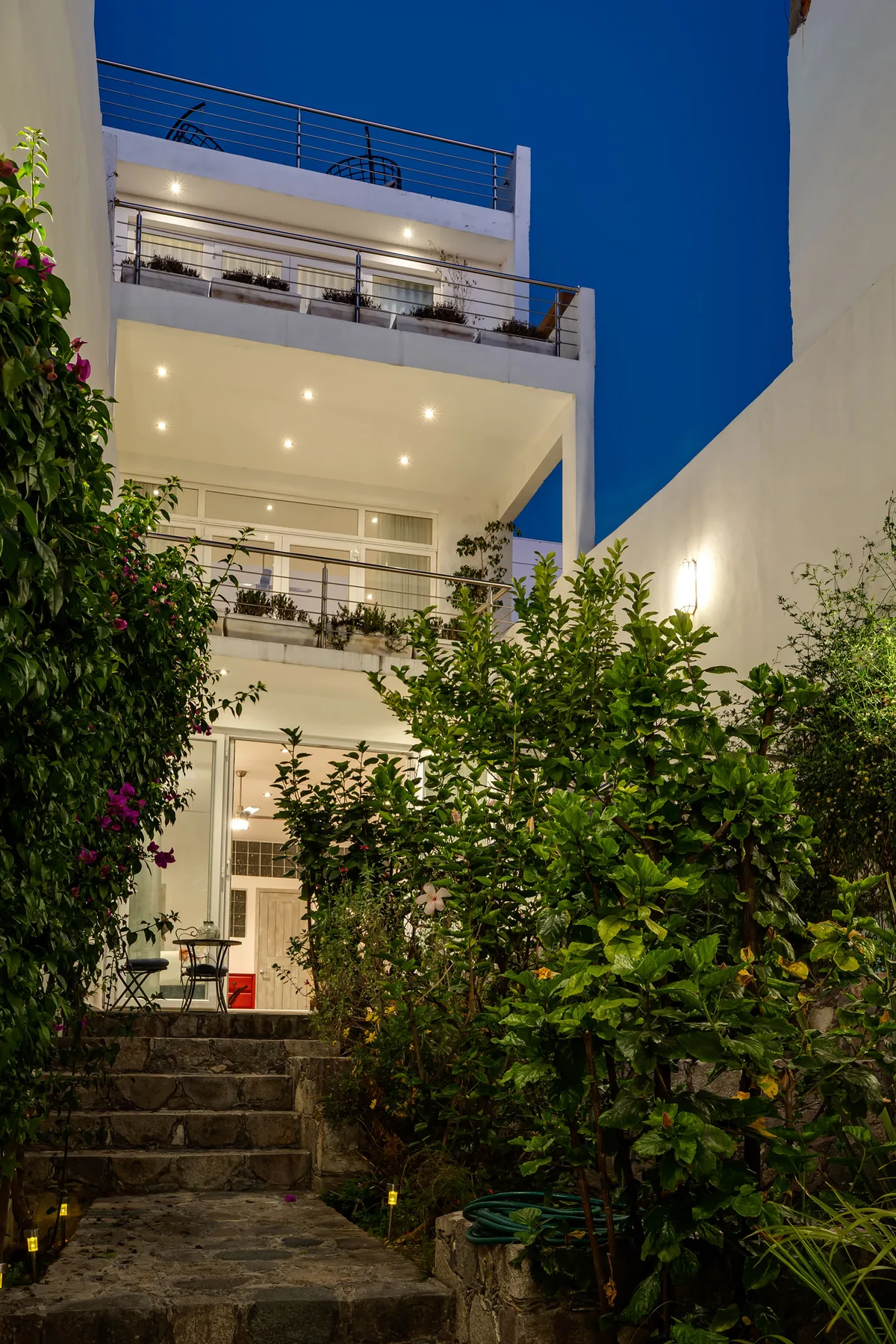 A white multi-level building stands behind a leafy garden and stone steps.
