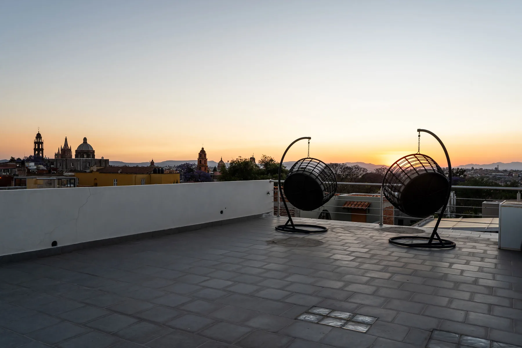 A rooftop terrace at sunset features two hanging seats and a city skyline.