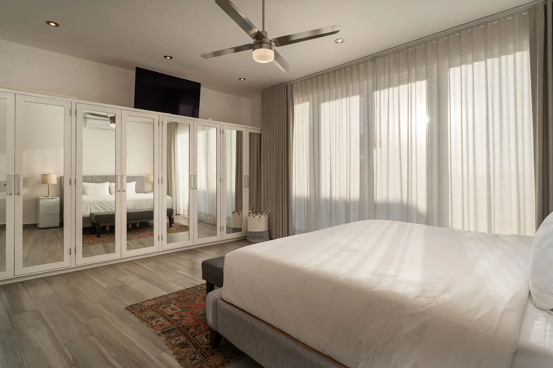 A bright bedroom with a large bed, mirrored wardrobes, and sheer curtains.