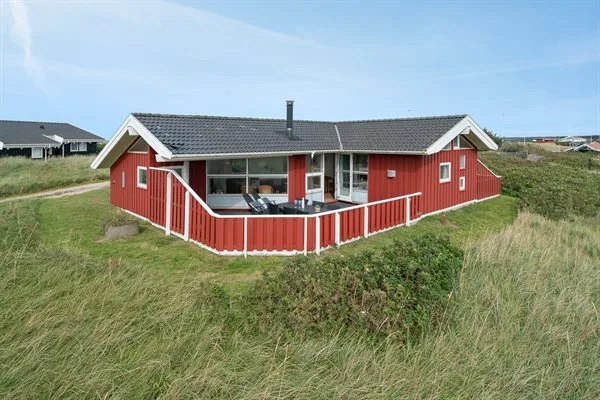 Una casa roja con una terraza de barandilla blanca está entre dunas cubiertas de hierba.