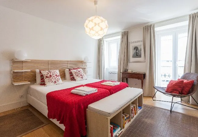 A bright bedroom features a bed with a red cover and a cushioned bench.