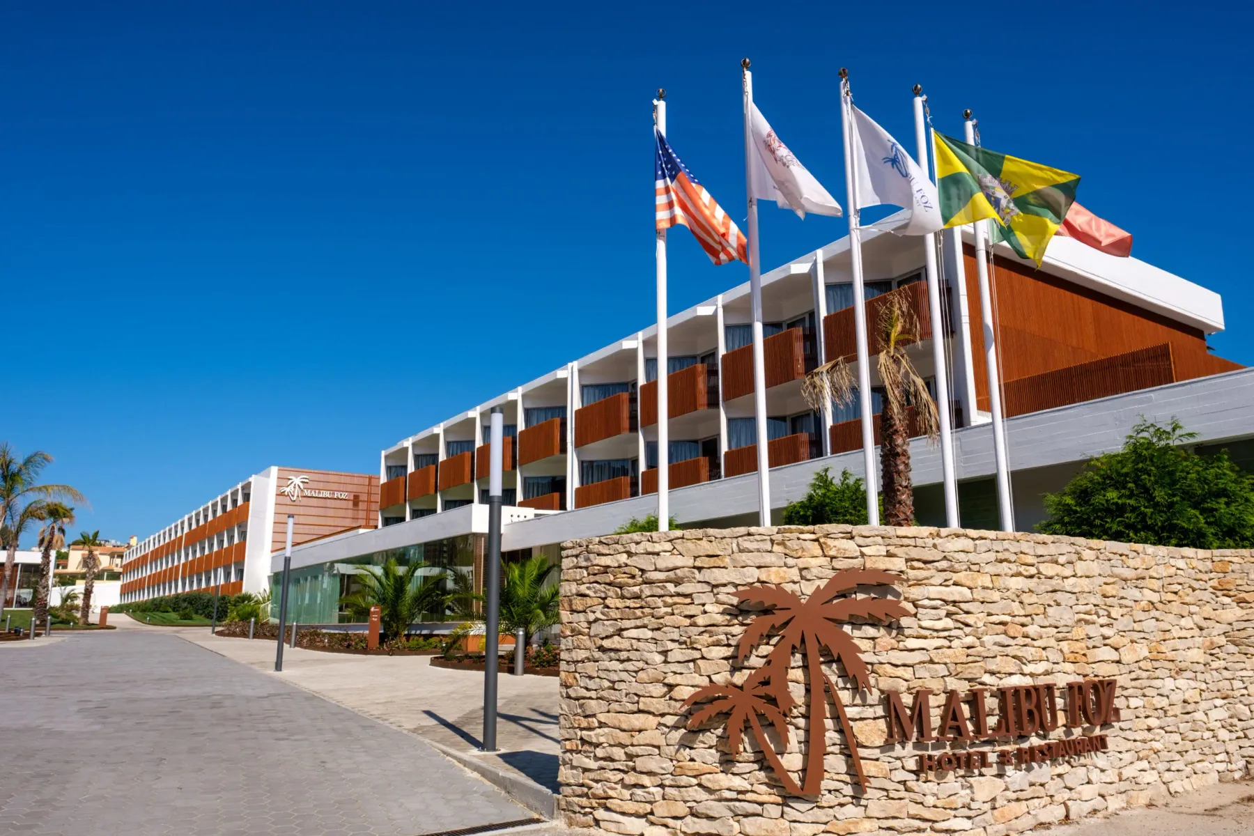 Espacio representativo del hotel: El exterior de un hotel moderno aparece tras un letrero de piedra y varias banderas.