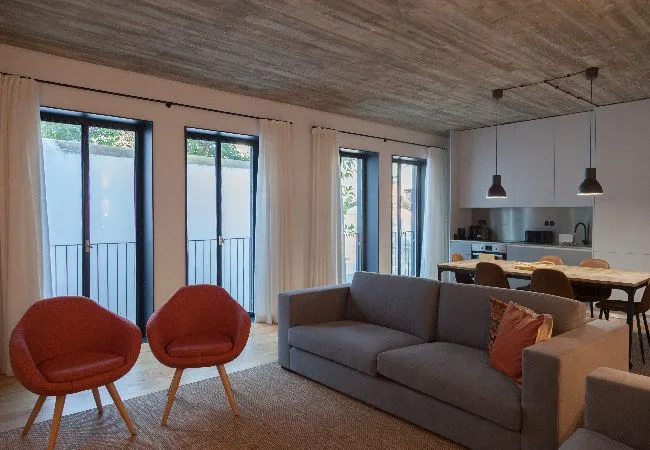 Una sala de estar y comedor con un sofá gris, sillones rojos y ventanas grandes.