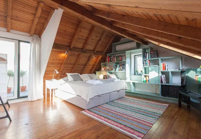 A bright attic bedroom with a bed, exposed wooden ceiling, and built-in shelving.