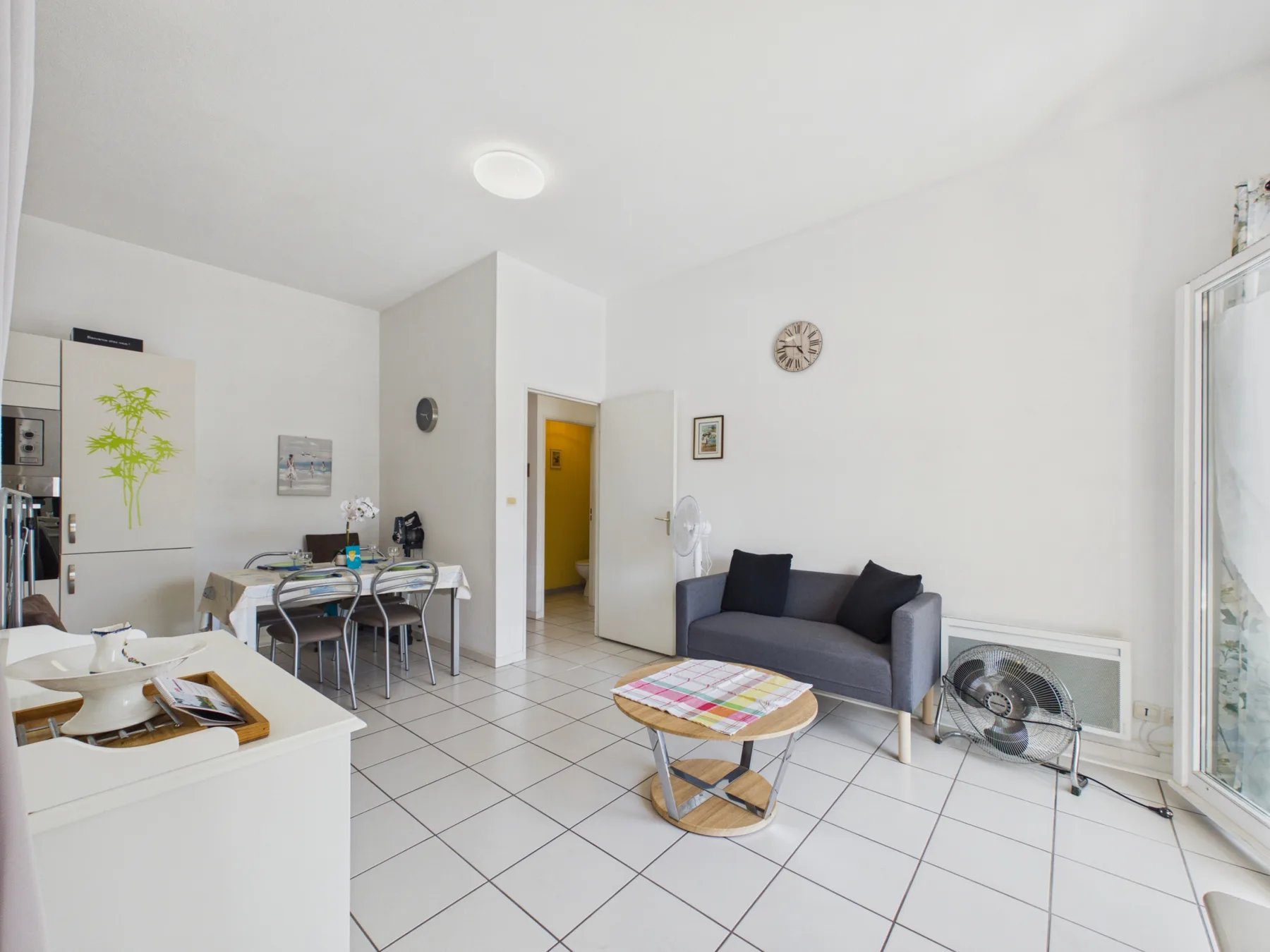 A bright open-plan living and dining area with a sofa, table, and white tiled floor.
