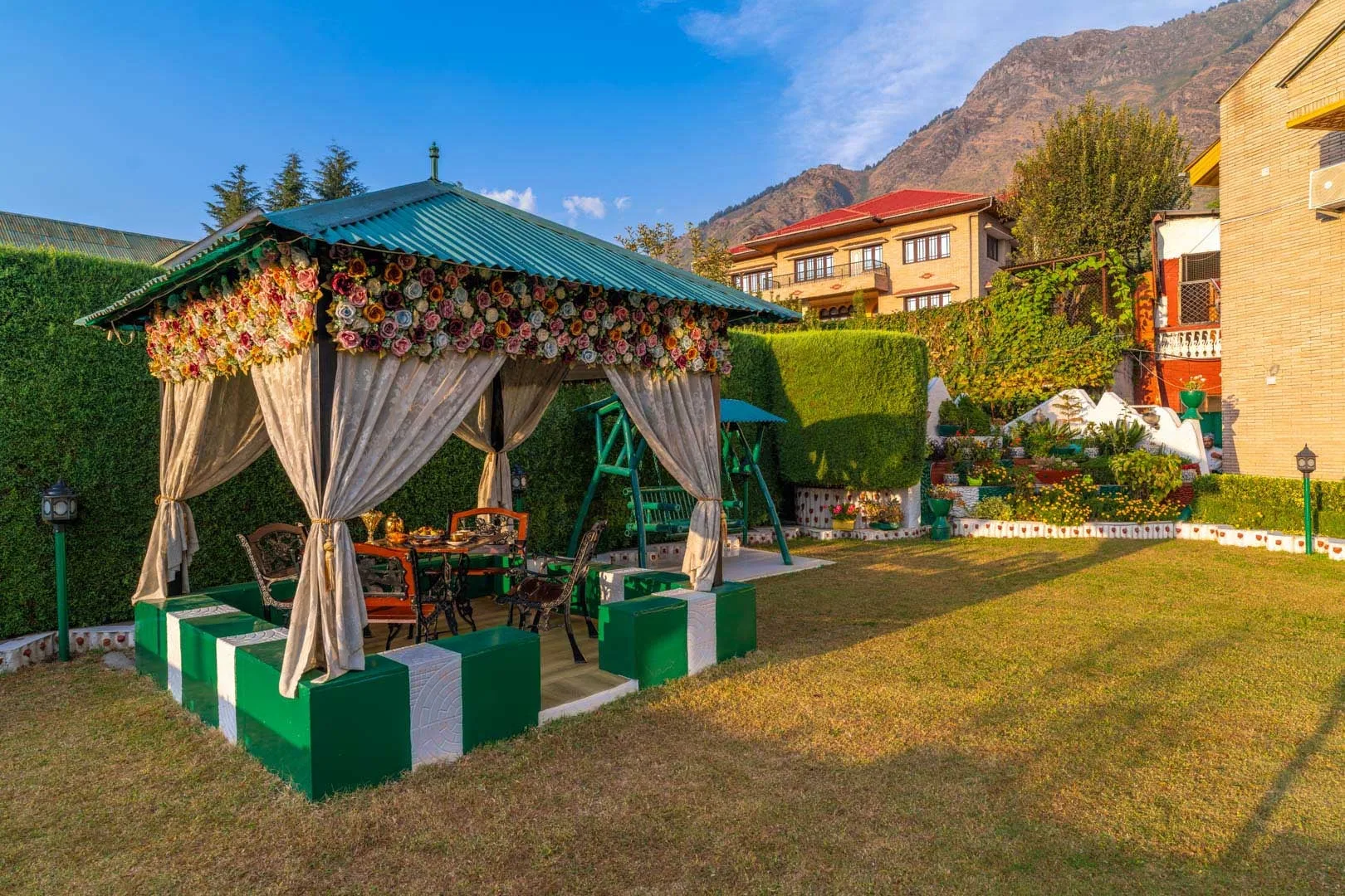 A garden seating pavilion with draped curtains and a trimmed hedge.