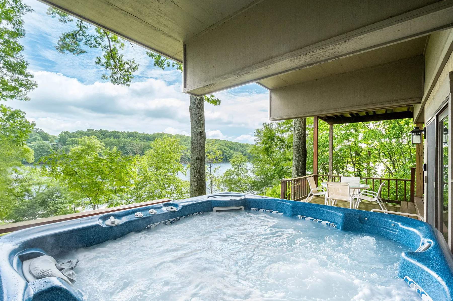 A bubbling outdoor hot tub sits on a covered deck overlooking a lake and trees.