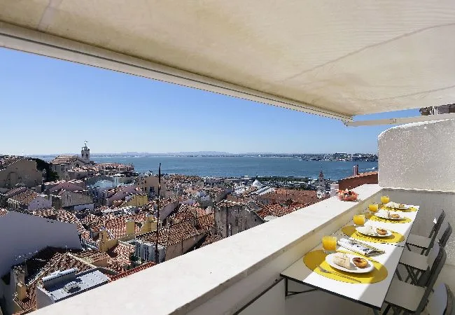 A rooftop terrace overlooks a coastal city and bay, with a dining table beneath an awning.