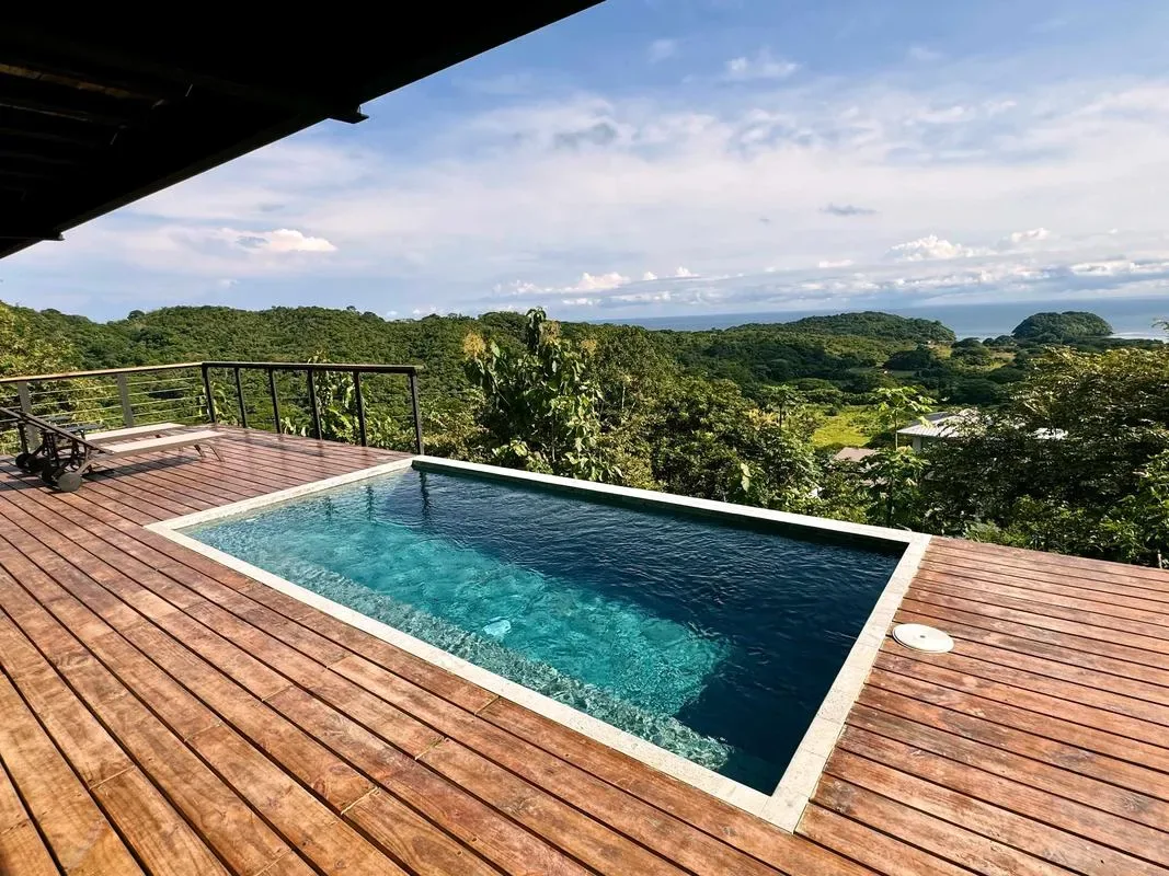 An outdoor swimming pool on a wooden deck overlooks a green, tree-covered landscape.