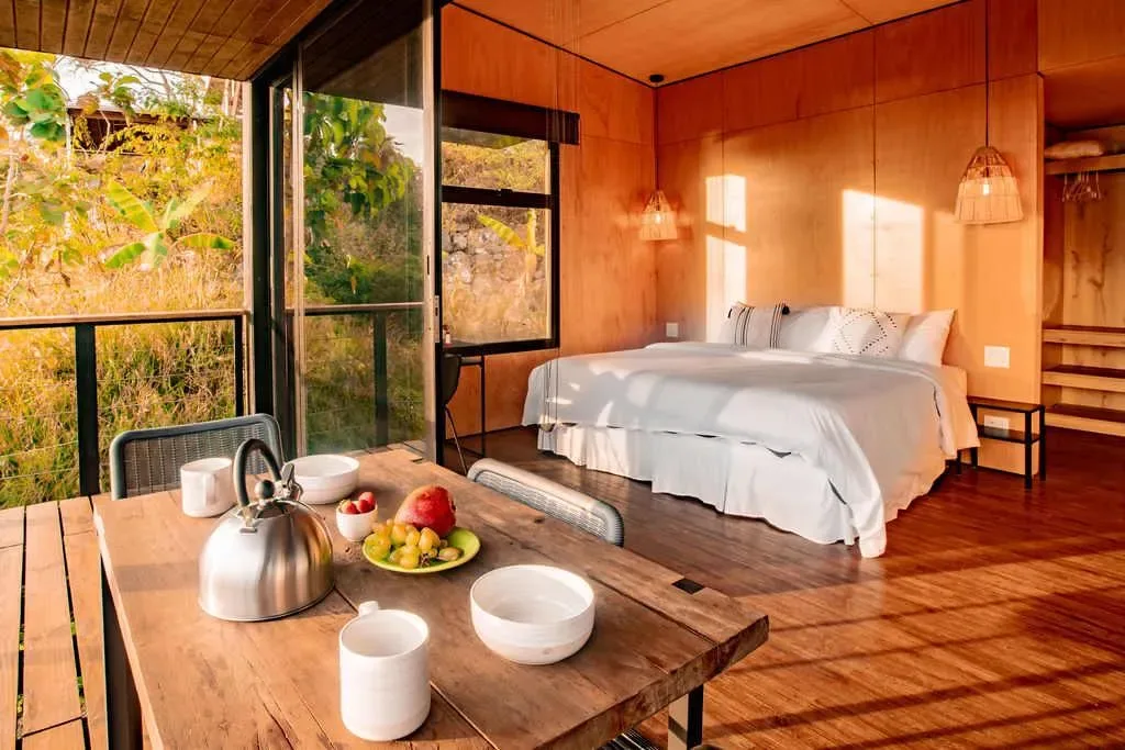 A bedroom with a white bed beside large windows overlooking greenery.
