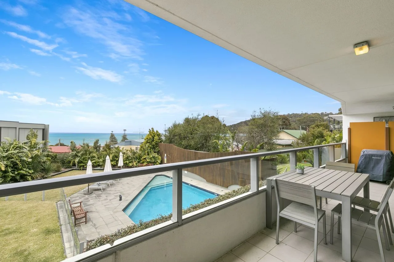 A covered balcony overlooks a swimming pool, greenery, and the sea.