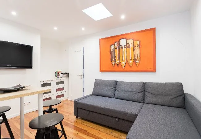 Una sala luminosa con asientos grises, un televisor en la pared y arte colorido.
