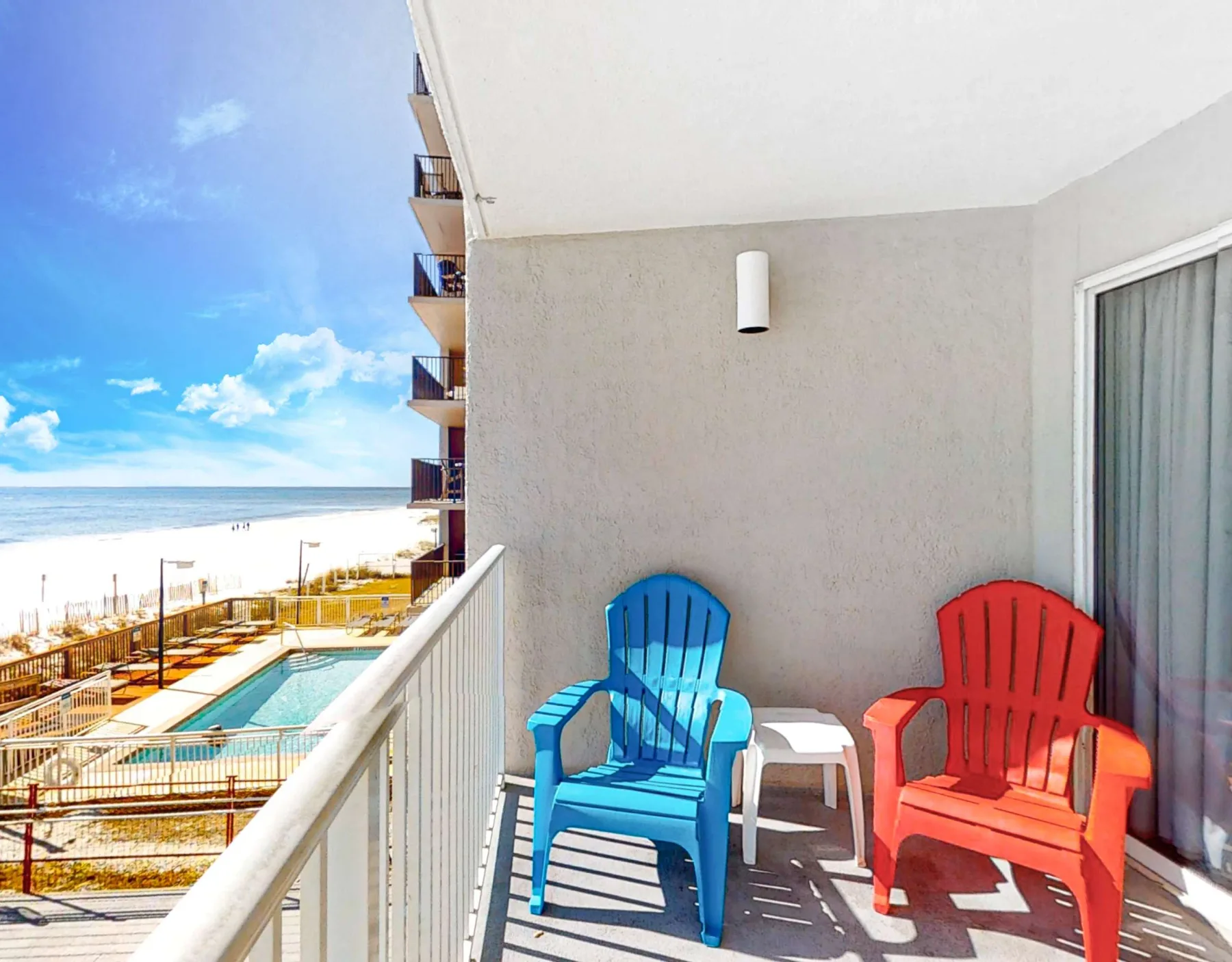A balcony with blue and red seating overlooks a pool and the ocean.