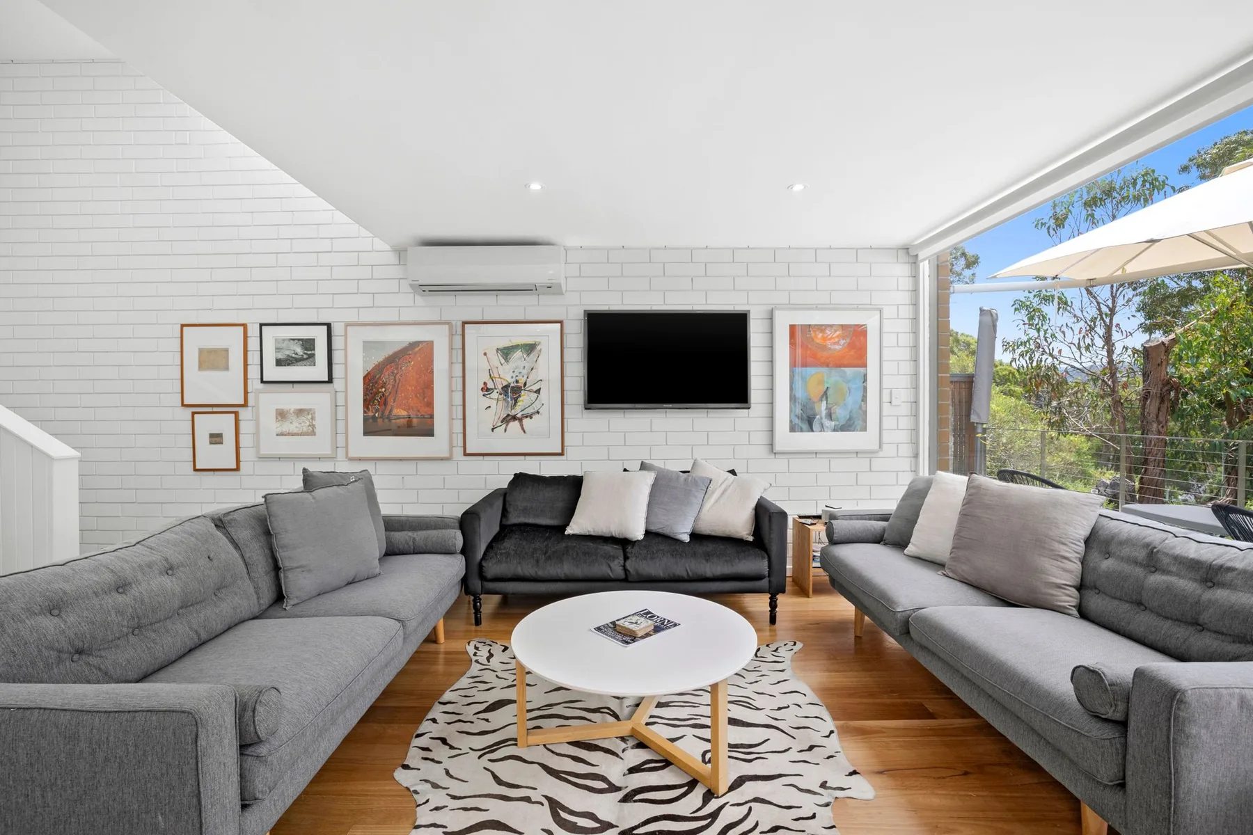 A bright living room with gray seating, a wall-mounted television, and large windows.