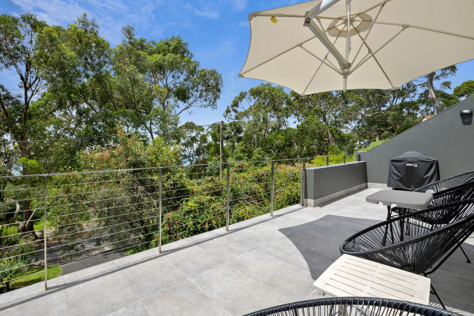 An outdoor terrace with seating, a large umbrella, and leafy trees beyond the railing.