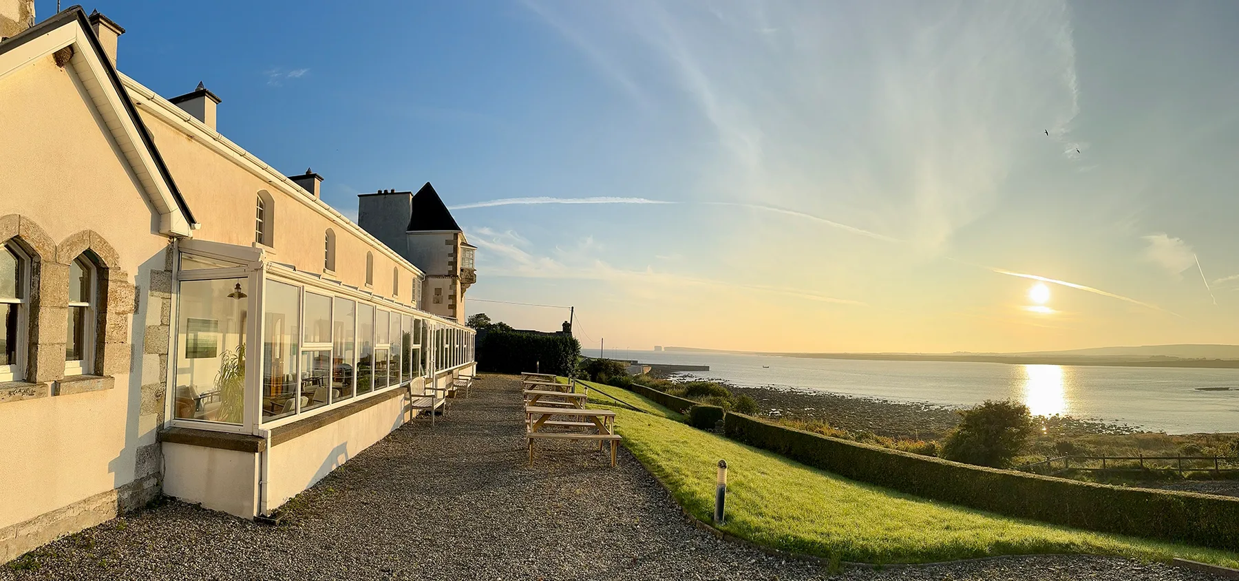 Representative hotel space: A coastal building beside a gravel path and grassy shoreline at sunset.