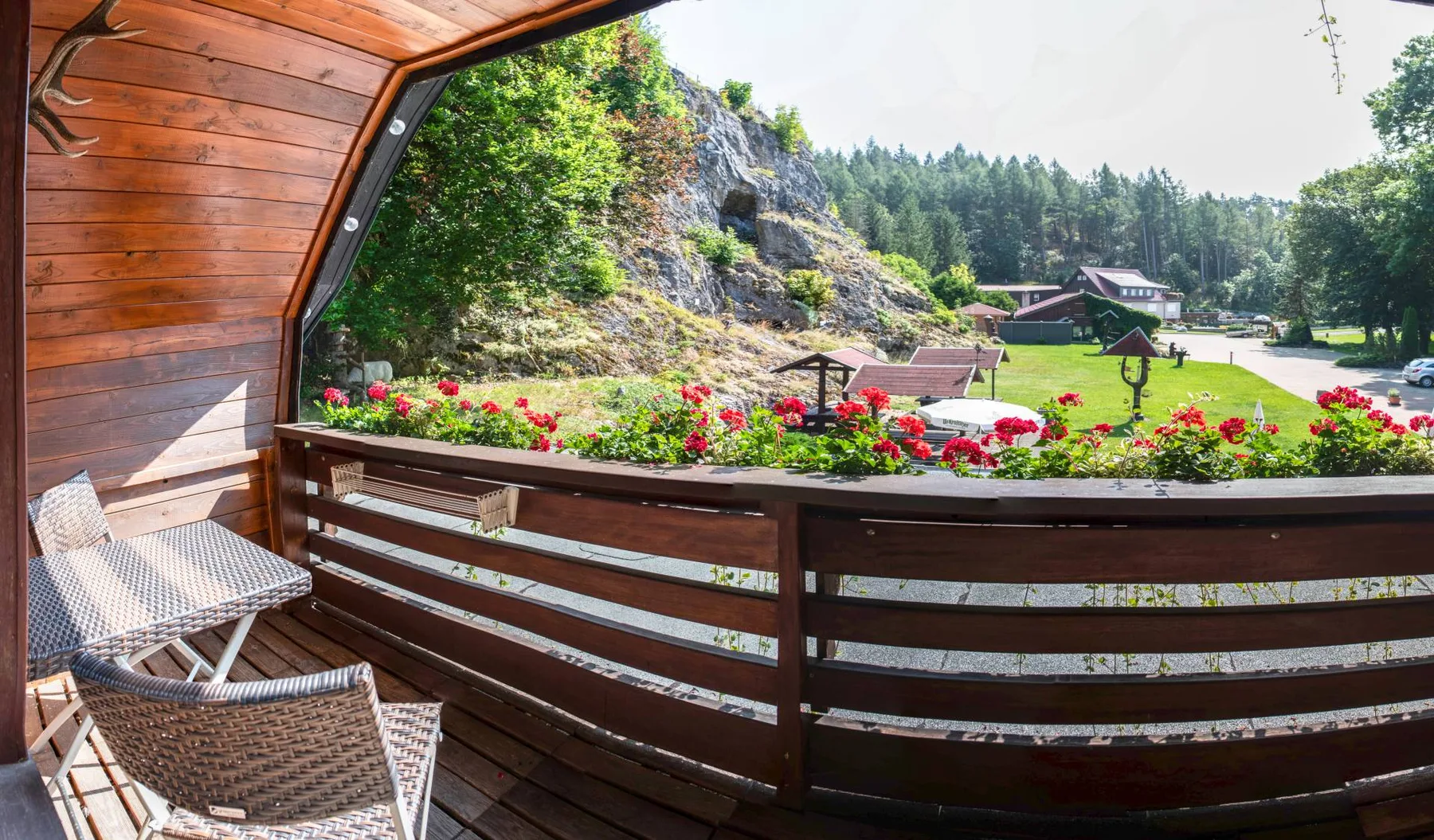 Espacio representativo del hotel: Un balcón de madera da a jardines floridos, una ladera rocosa y un paisaje boscoso.
