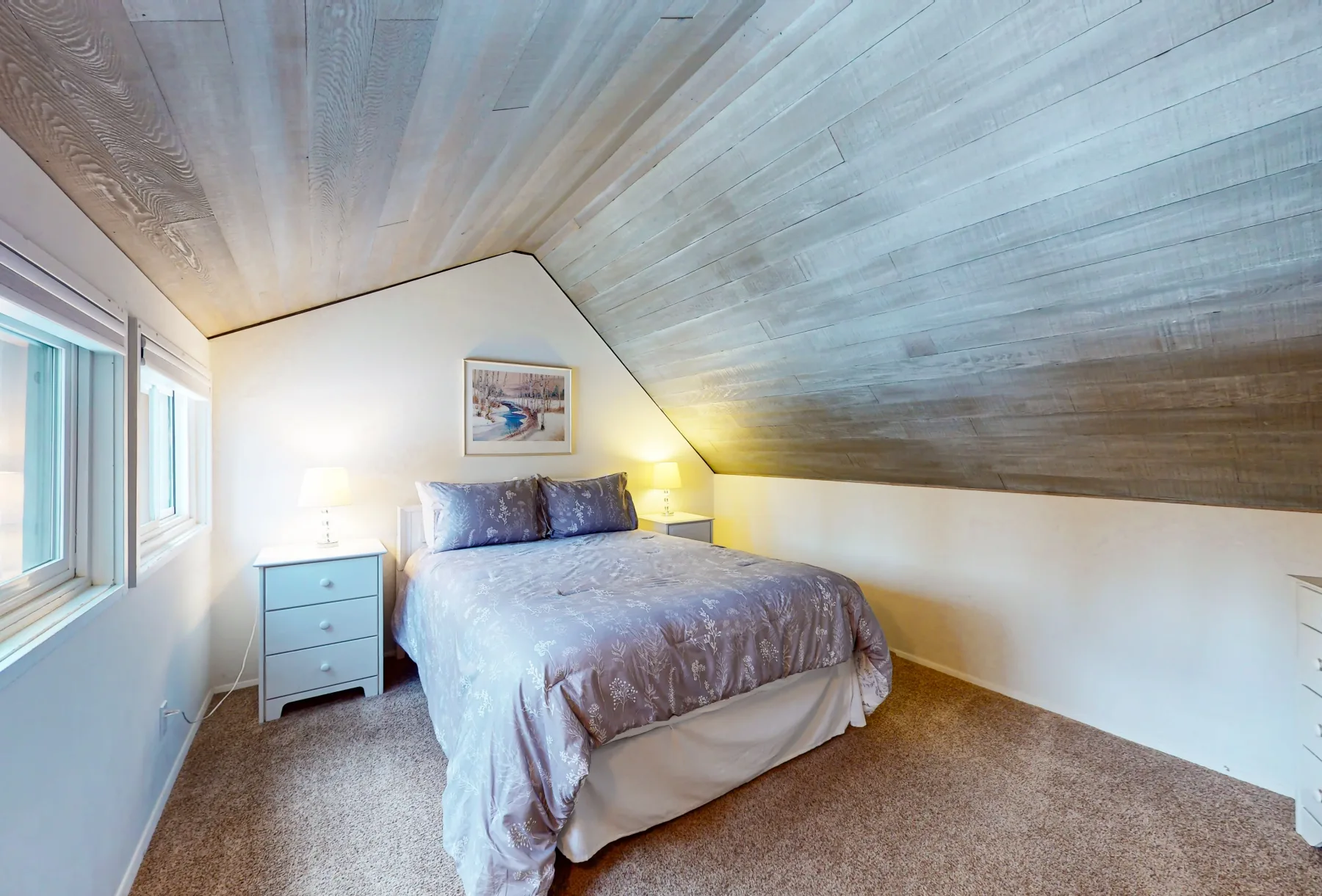 Un dormitorio con una cama bajo un techo inclinado y ventanas junto a una pared.