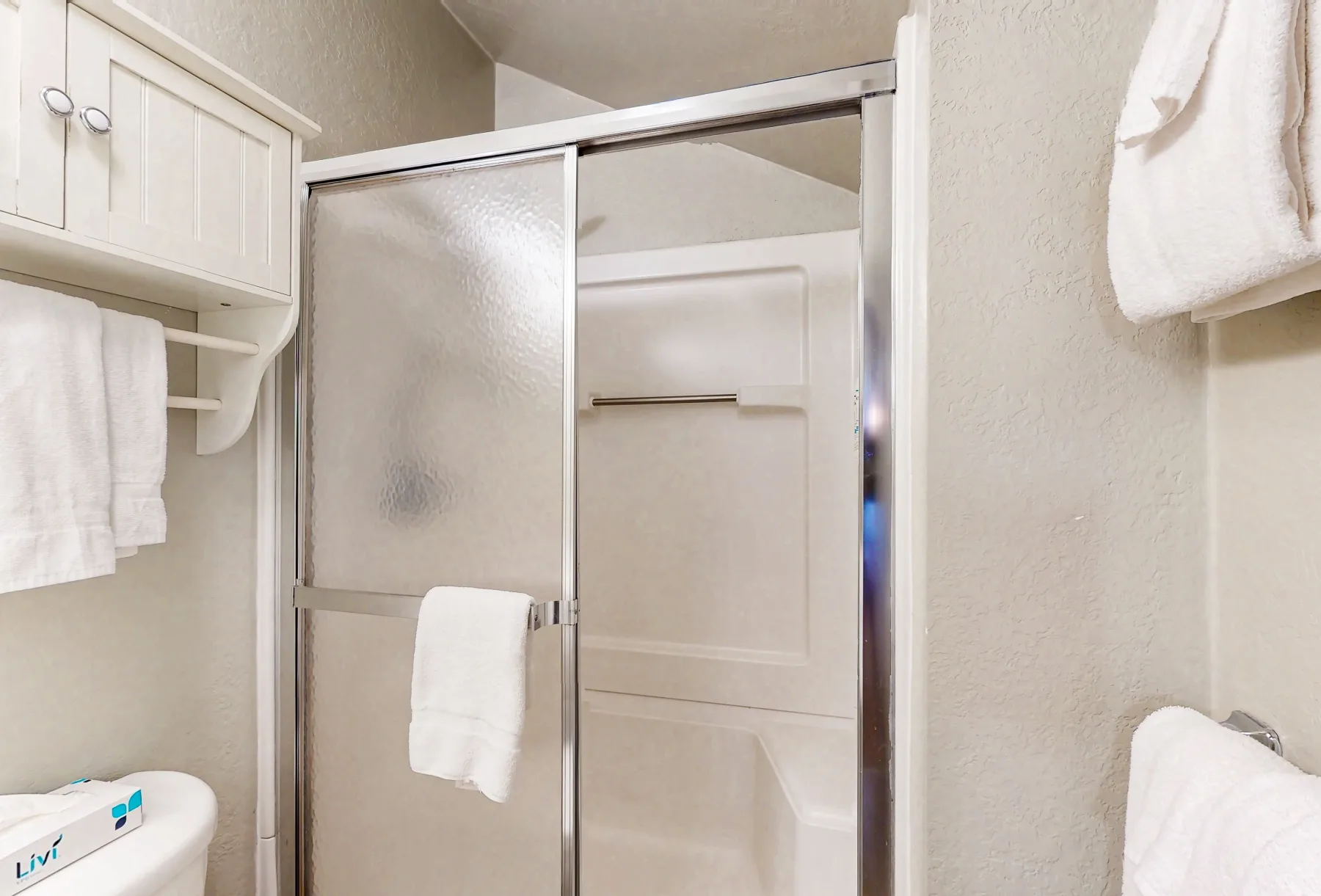 Zona de ducha en un baño con puerta corrediza de vidrio y toallas blancas.