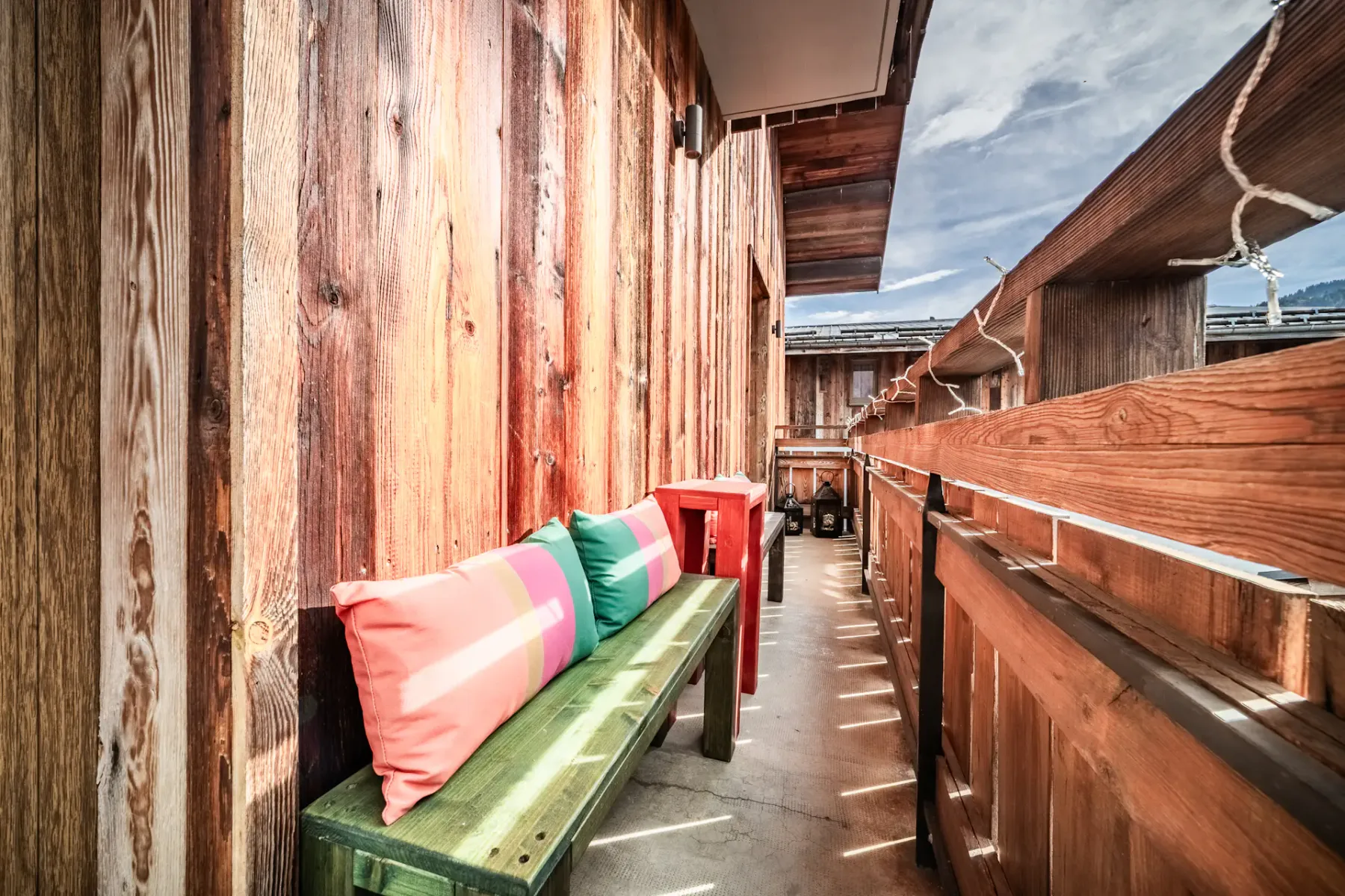 A narrow wooden balcony walkway has a green bench with colorful cushions.