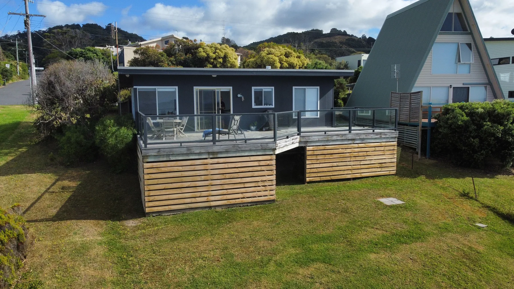 A house with a raised deck and glass railing faces a grassy yard.