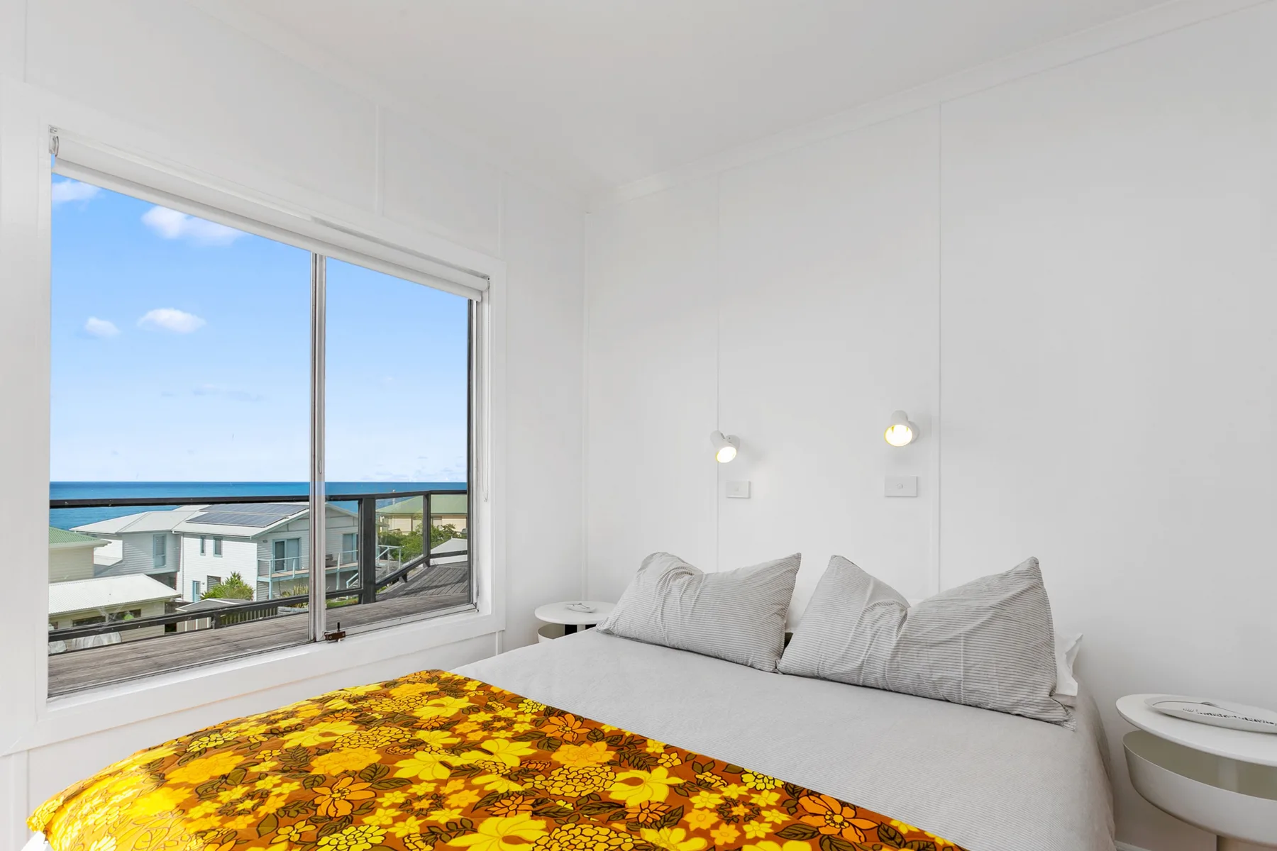 A bright bedroom with a large window overlooking the ocean and a colorful bedspread.