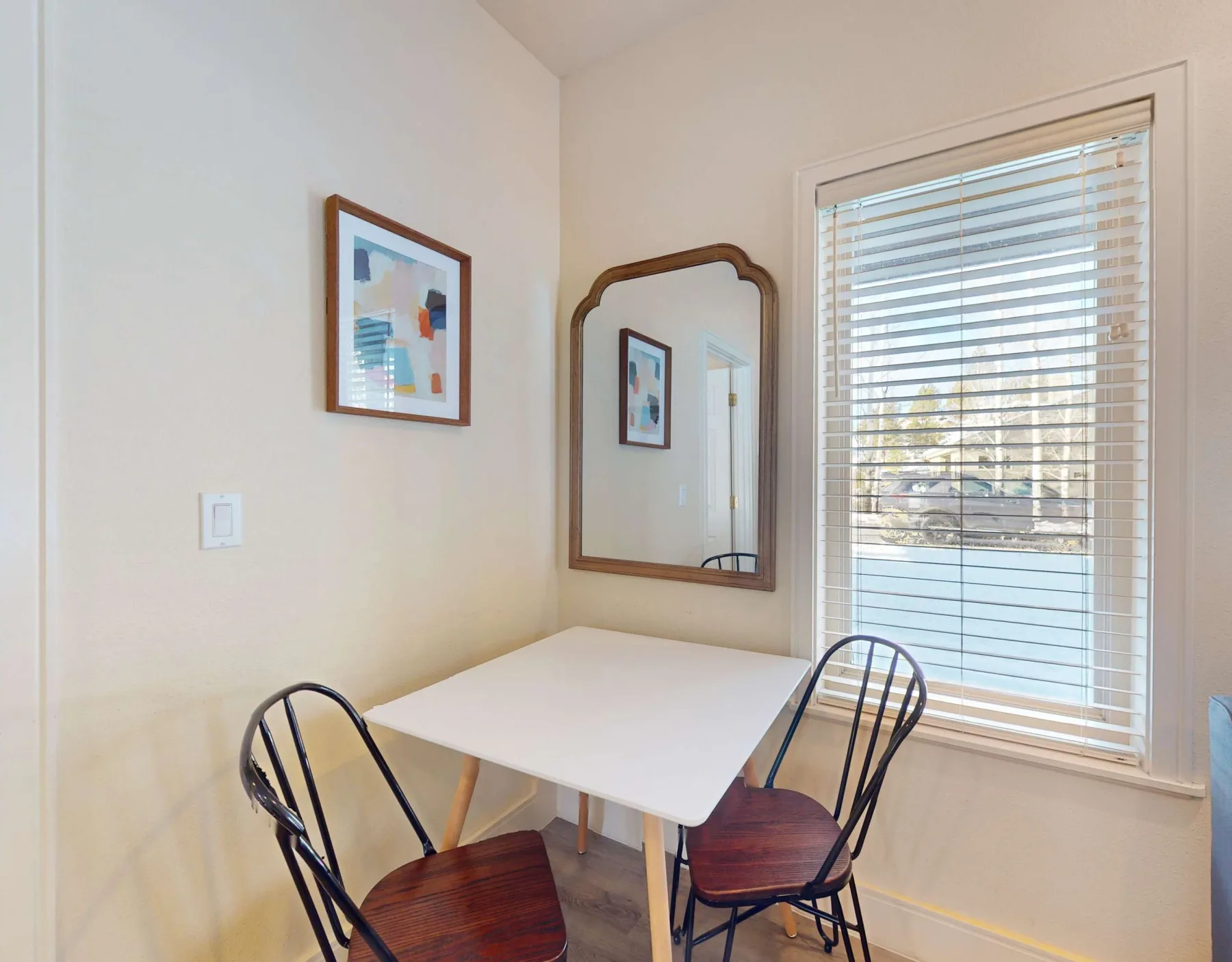 Un rincón de comedor luminoso con una mesa blanca, asientos oscuros y una ventana grande.