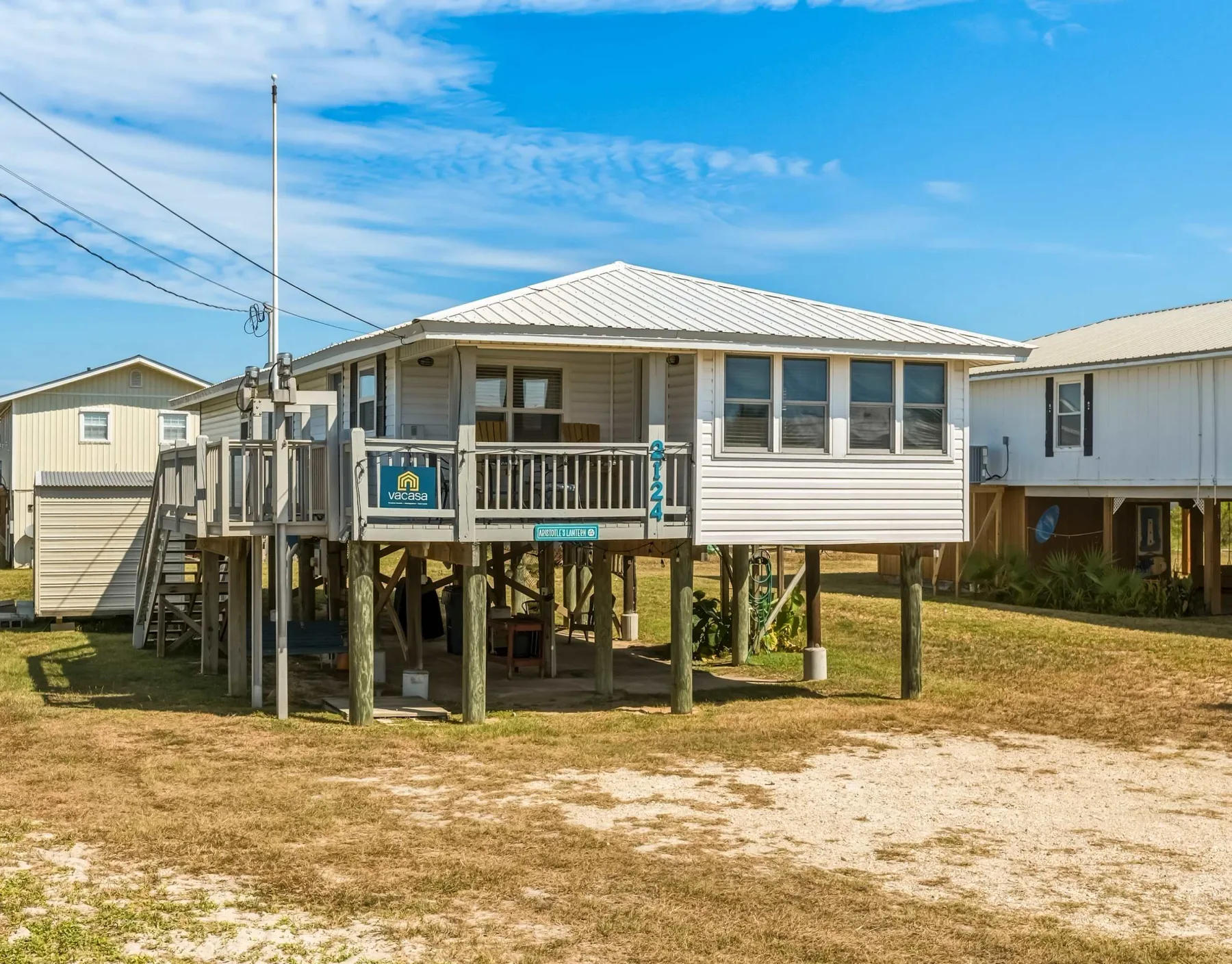 A raised coastal-style house stands on supports above a sandy yard.