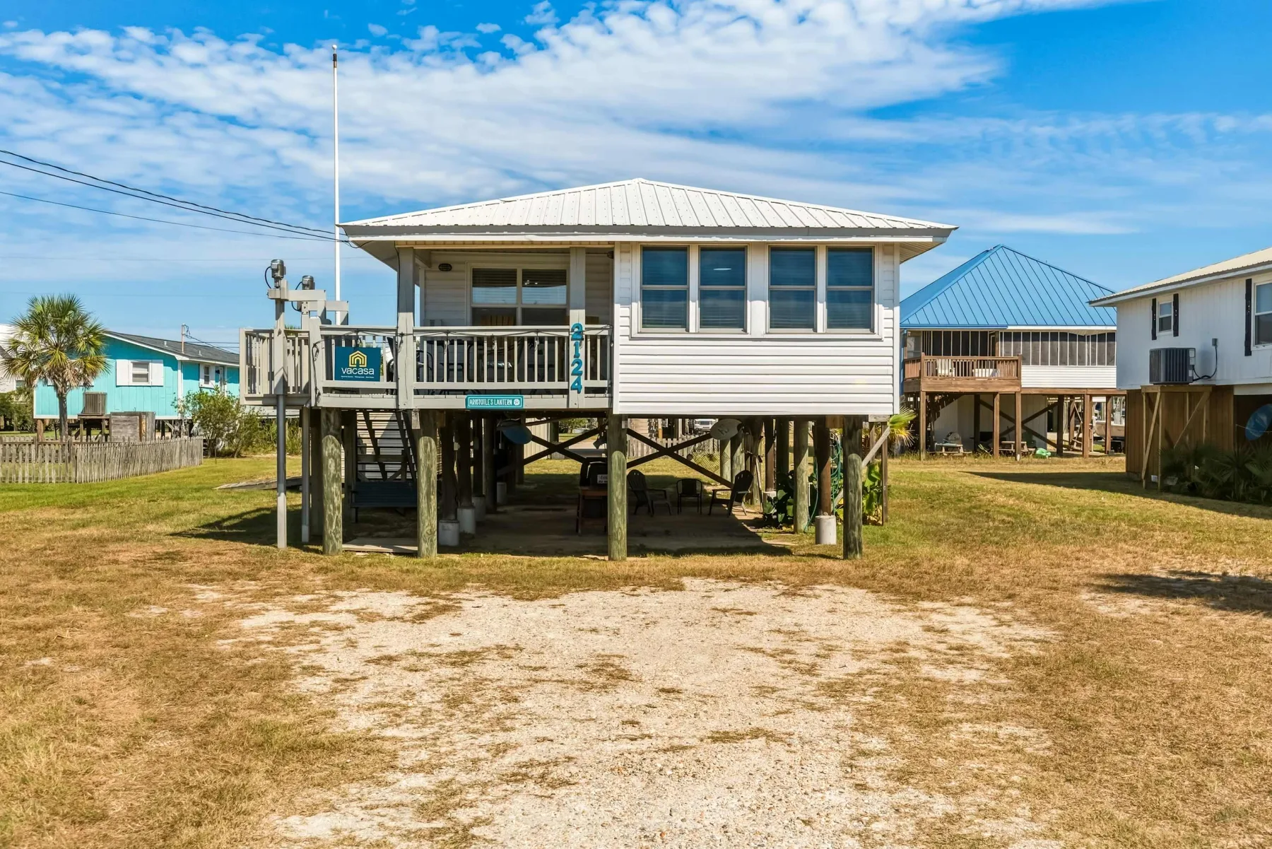 A raised coastal house stands above a sandy yard beneath a blue sky.
