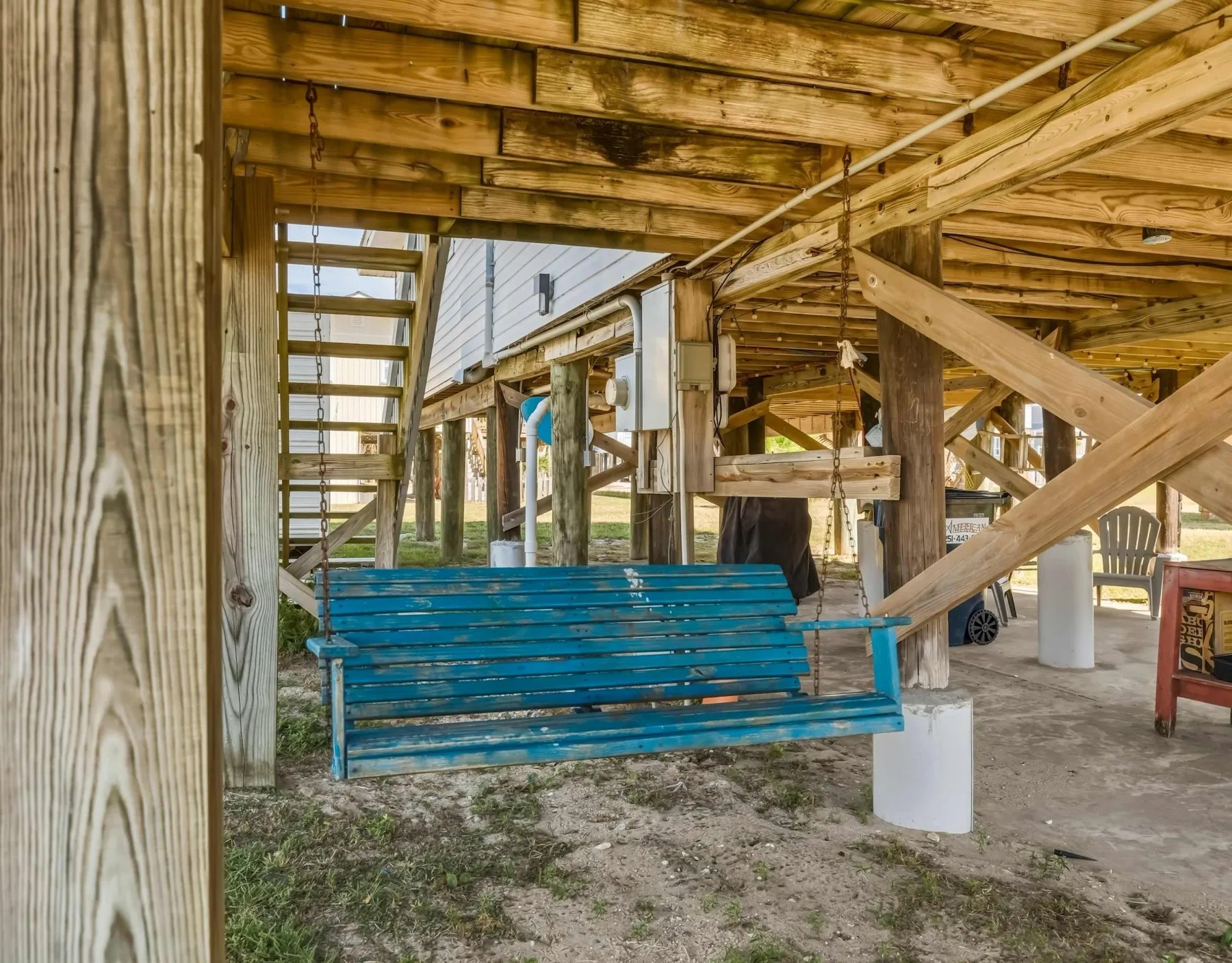 A blue swing hangs beneath a wooden raised structure beside a parked car.