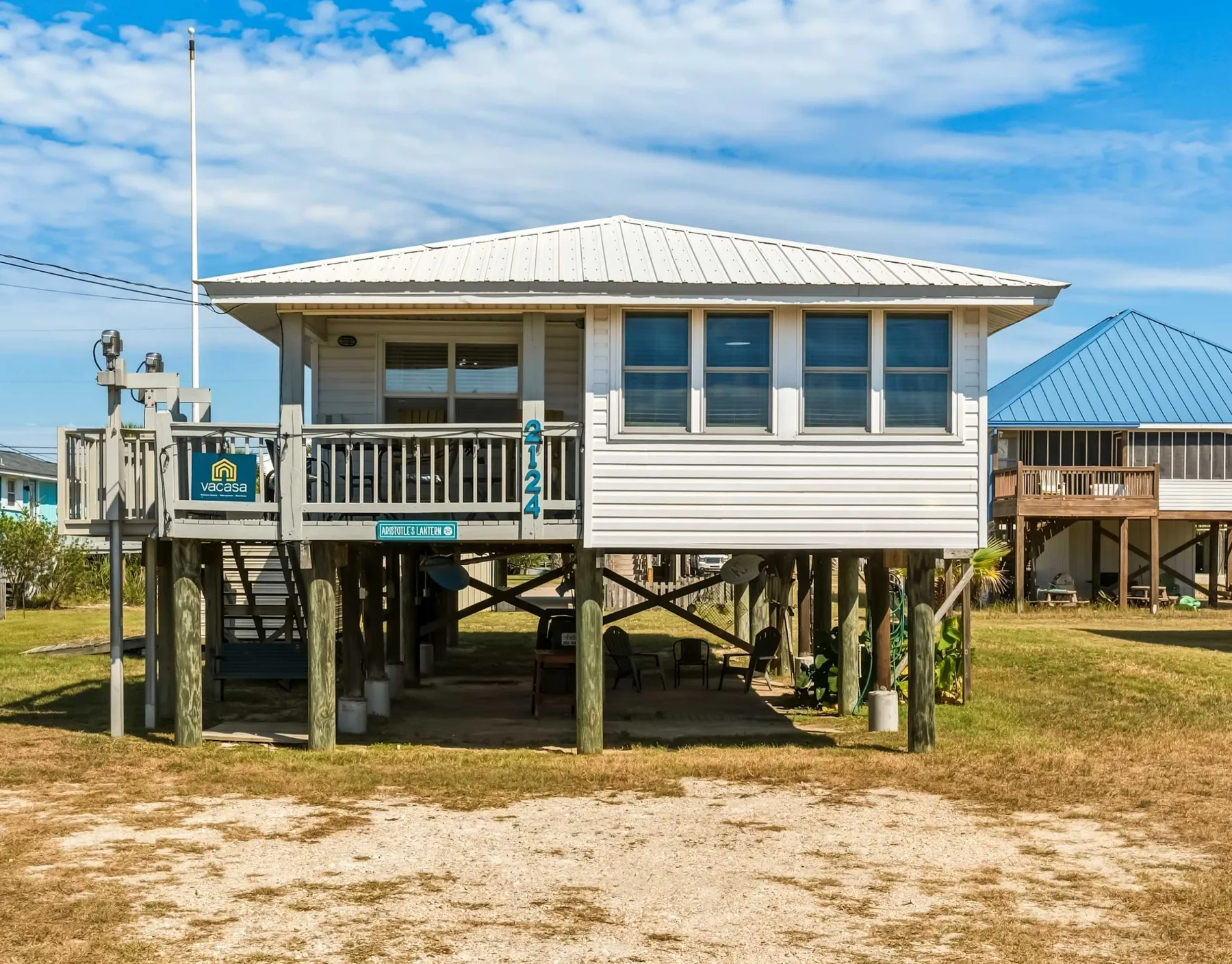 A raised coastal house with a covered balcony stands above a grassy lot.