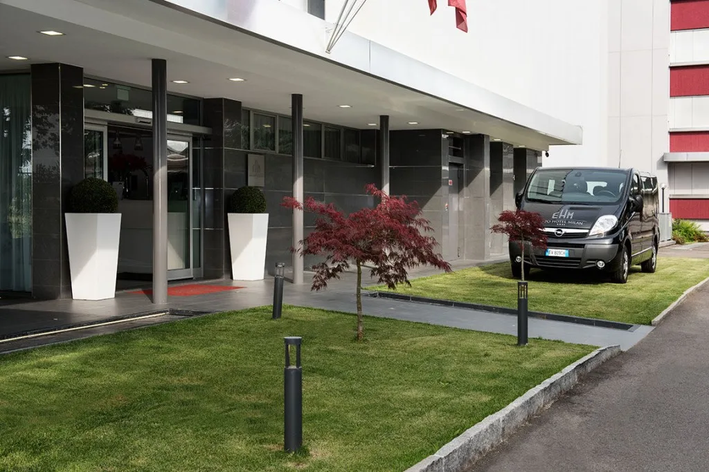 Espacio representativo del hotel: Entrada de hotel con puertas de vidrio, césped ajardinado y una furgoneta estacionada.