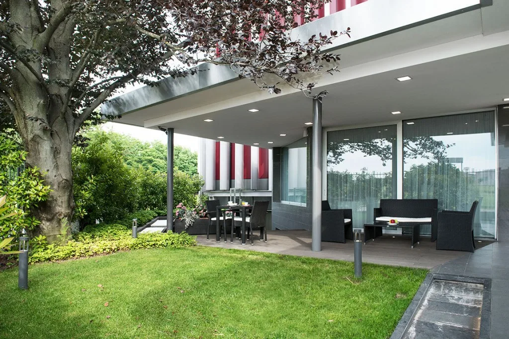 Espacio representativo del hotel: Una zona exterior cubierta con asientos junto a un césped y grandes ventanas.