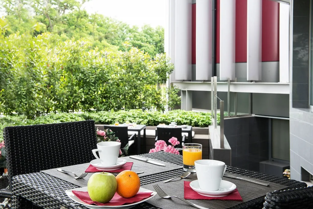 Espacio representativo del hotel: Una mesa de comedor al aire libre tiene tazas, zumo y fruta junto a plantas frondosas.