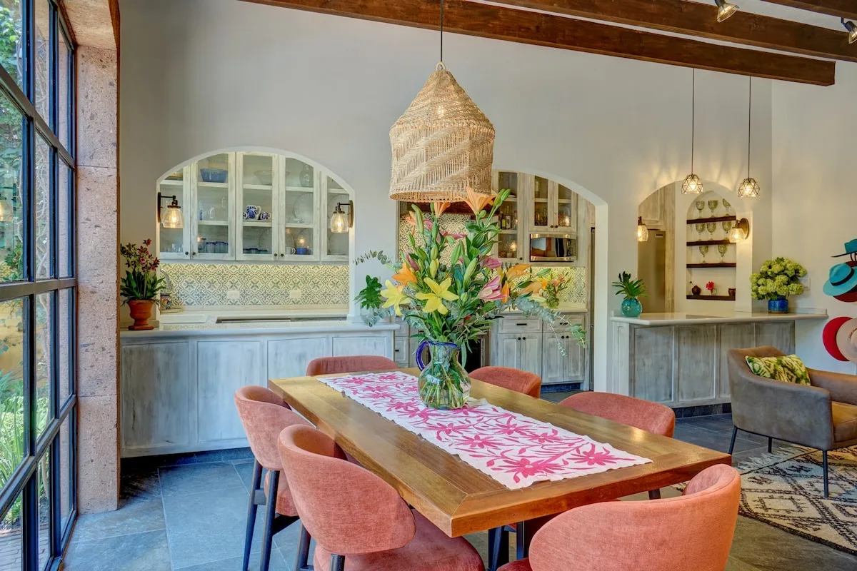 Un comedor con una mesa de madera, asientos rosas y un centro floral.