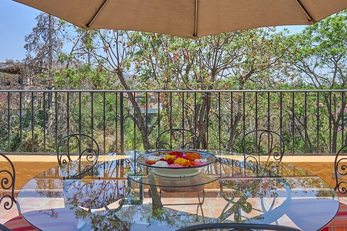 Una mesa exterior con superficie de vidrio y asientos está bajo una sombrilla en una terraza arbolada.
