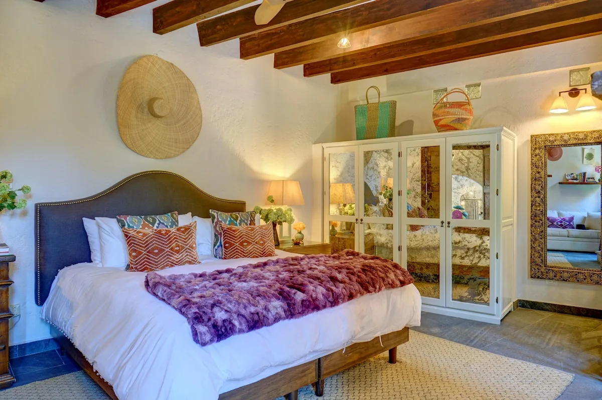 Un dormitorio con una cama, cojines estampados y un armario con espejo bajo vigas de madera.