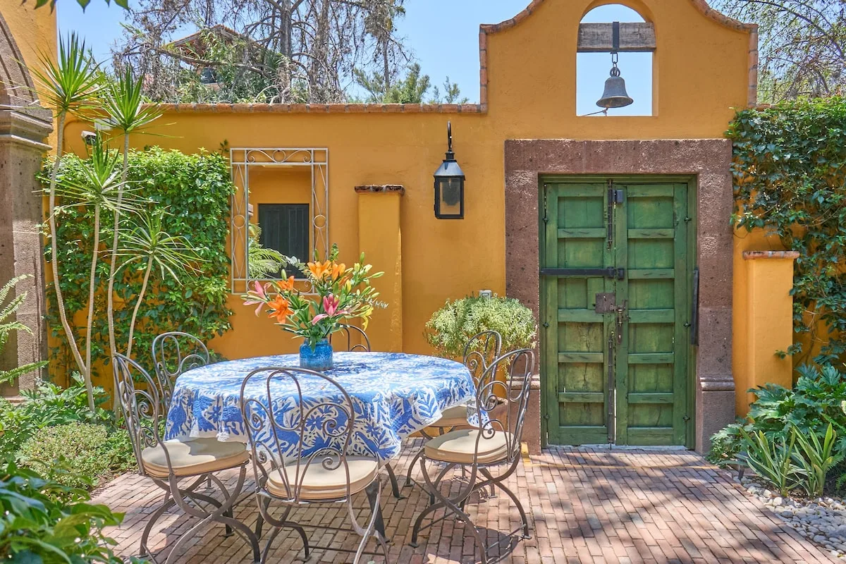 Un patio al aire libre con una mesa redonda, sillas y una puerta verde en una pared ocre.
